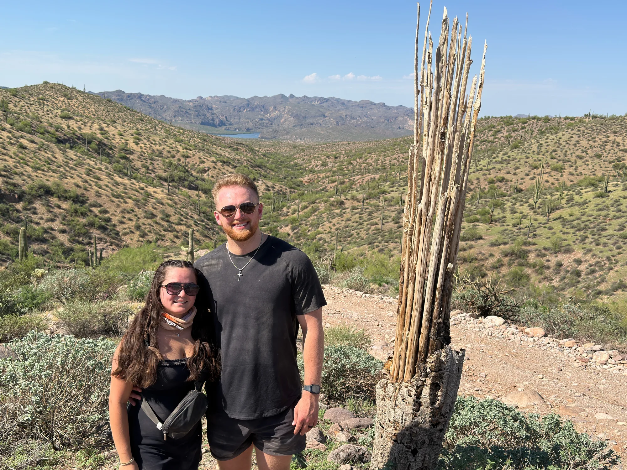 A couple standing beside a weathered saguaro skeleton on a desert hillside, with a lake and mountains in the distance east of Scottsdale